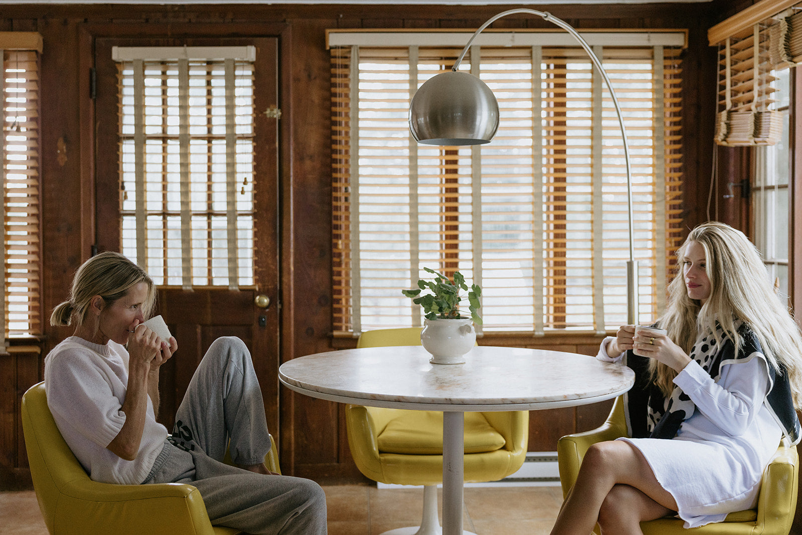 Two women sit in yellow chairs at a round table with a potted plant, drinking from cups in a cozy, wood-paneled room with large windows and blinds.