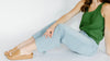 Woman in green tank top and blue jeans sitting with one leg bent, wearing tan slide sandals against a white background.