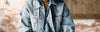 Person wearing a blue denim jacket with their hands folded, standing in front of a blurred brick and wood wall background.