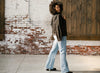 A person with curly hair wearing a black jacket and light blue jeans is standing in front of a brick and wooden wall, looking back over their shoulder.
