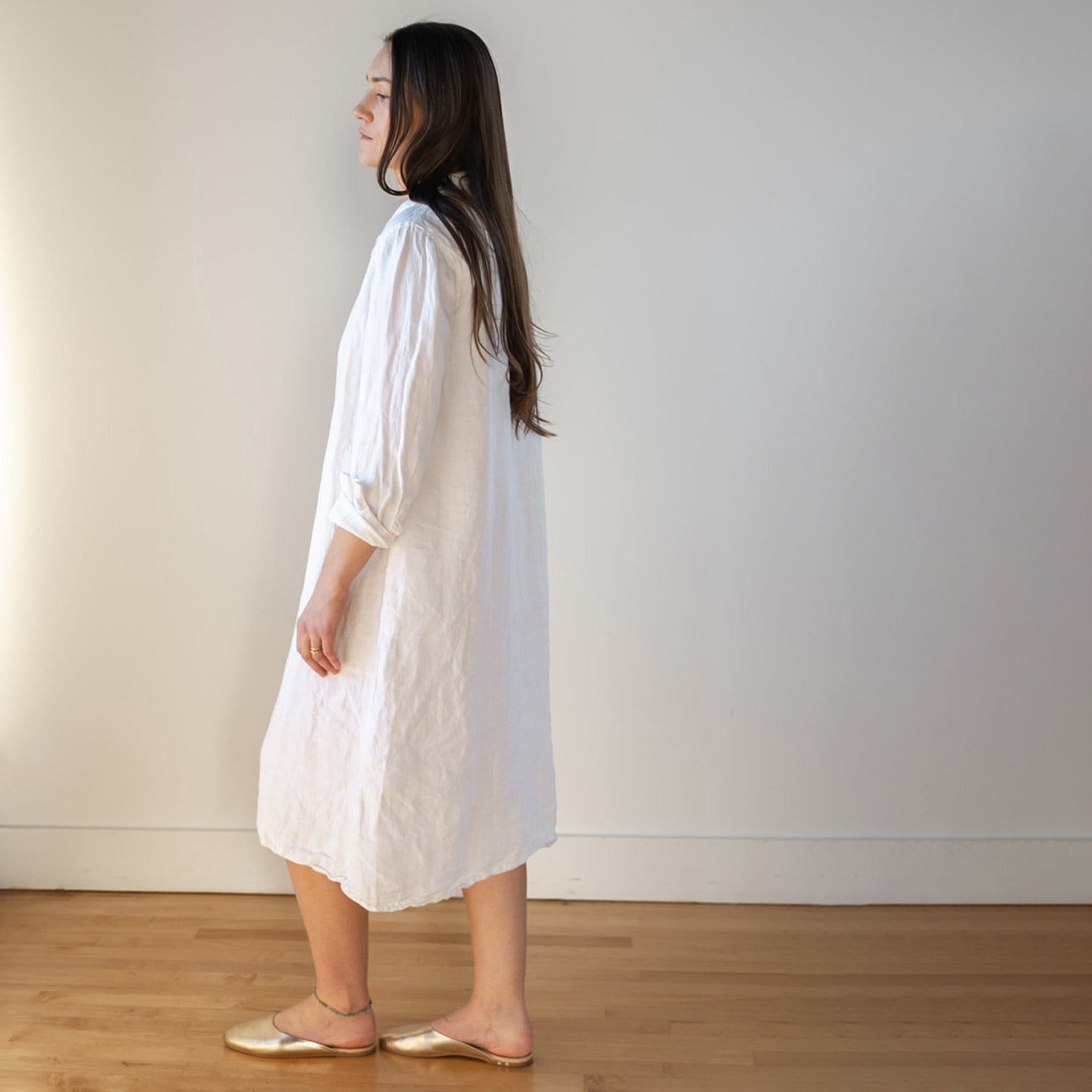 A woman with long brown hair stands in profile, wearing a loose, knee-length white dress and gold flat shoes, in a bright room with wooden floors and a plain white wall.