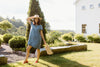 A woman in a blue dress and straw hat stands outdoors, smiling and holding a woven handbag. She poses near a landscaped garden with a white building and trees in the background.