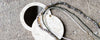Two circular, mother-of-pearl compact mirrors are on a textured gray surface. Several beaded necklaces in shades of gray, white, and black are draped across them.