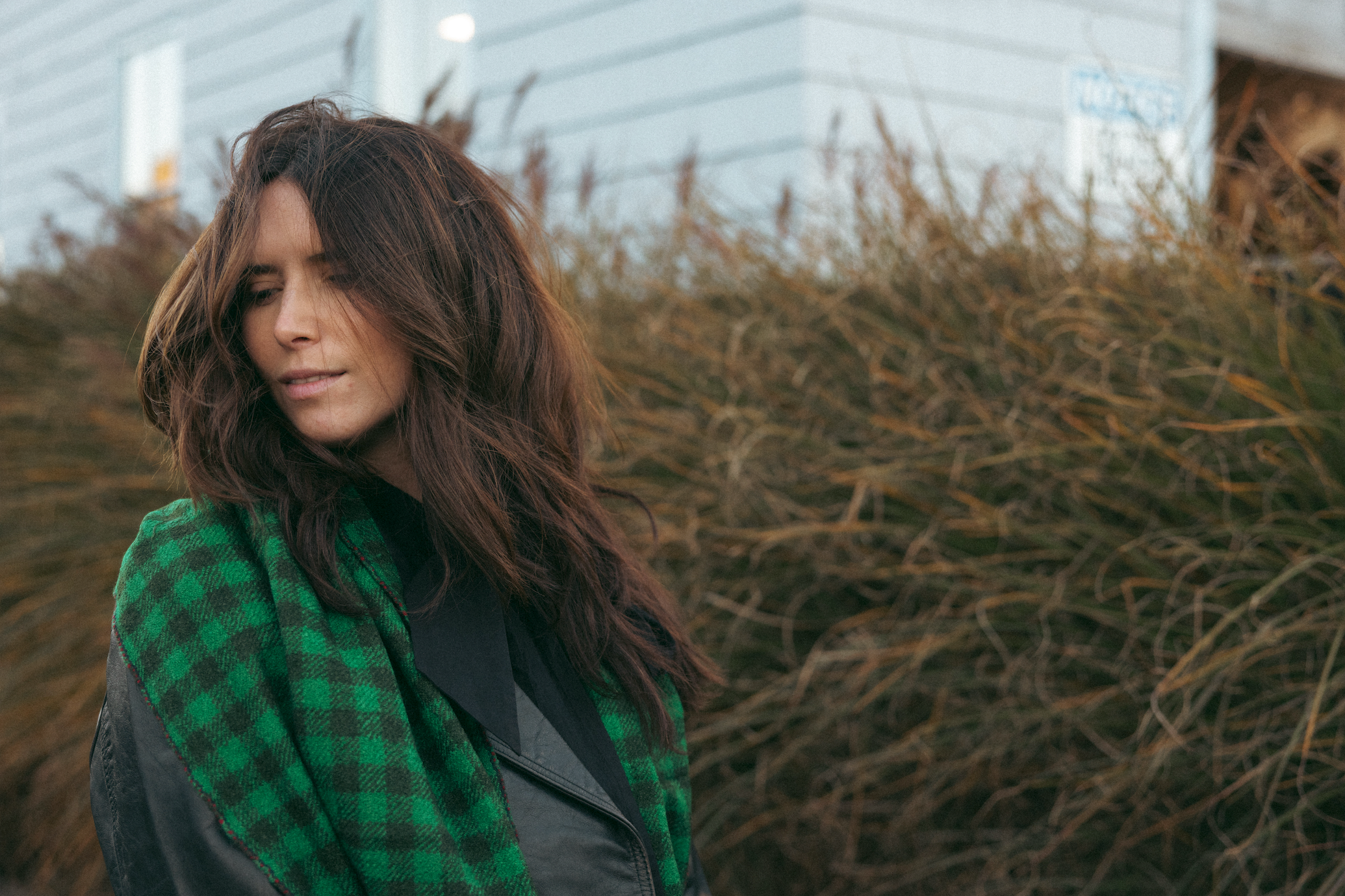 A woman with long brown hair stands outside, wearing a green and black checkered scarf and a dark jacket, with tall grasses and a building in the background.