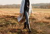 Person walking in a grassy field, wearing a long gray coat, black top, gray jeans, and black boots. Trees and a light sky form the backdrop.