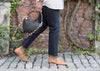 A person walks on a cobblestone path, wearing black jeans, tan shoes, and holding a black woven handbag. They are beside a brick wall covered with ivy and vines.
