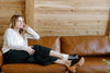 A person with long hair sits on a brown leather couch, wearing a white top, black pants, and wooden clogs, resting their head on their hand and looking to the side in a wood-paneled room.