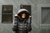 A person in a black puffer jacket and hood stands outdoors, smiling and pulling the hood over their head with both hands. There are windows and a door in the background.