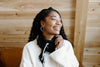 A woman with curly hair, wearing a white sweater and black scarf, smiles while looking to her left against a wooden interior background.