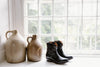 Two ceramic jugs and a pair of black leather boots are placed on a windowsill with a large window in the background.