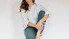 A smiling woman in a striped shirt and jeans sits casually on a stool, holding one knee with her hands.