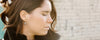 A woman with closed eyes, wearing pearl earrings and a black collared jacket, stands in front of a blurred brick wall.