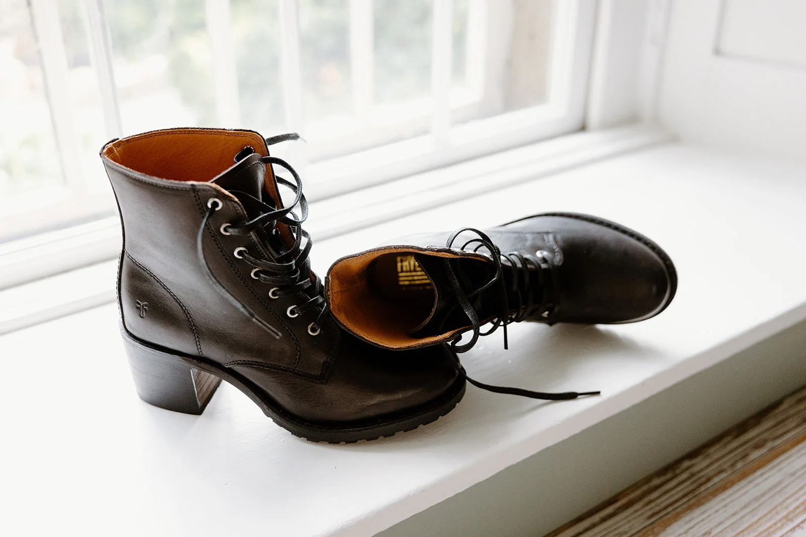 A pair of black lace-up leather ankle boots with a low chunky heel, placed on a white windowsill.