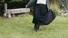 Person in a black dress standing in a grassy field, holding a brown fur handbag.