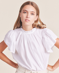 A woman wearing a Trovata Birds of Paradis Carla Highneck Shirt in White with ruffle sleeves stands against a neutral background.