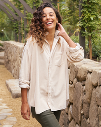 A woman with curly hair smiling outdoors while standing on a garden path, wearing the Frank & Eileen Shirley Oversized Button Up in Vintage White Cotton.