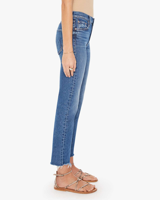Side view of a person standing and wearing blue Mother Denim The Mid Rise Rider Ankle Fray in Local Charm jeans with frayed hems and strappy sandals.