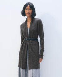 A woman with short dark hair wears the White + Warren Cashmere Trapeze Cardigan in Dark Slate Heather, belted at the waist over a light gray dress, standing against a plain light background.
