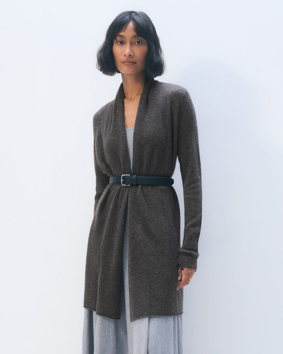 A woman with short dark hair wears the White + Warren Cashmere Trapeze Cardigan in Dark Slate Heather, belted at the waist over a light gray dress, standing against a plain light background.