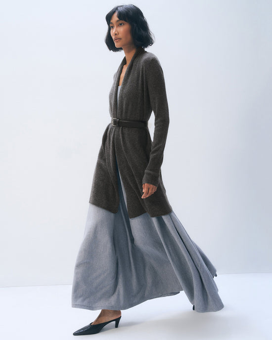A woman with a bob haircut walks in profile, wearing the White + Warren Cashmere Trapeze Cardigan in Dark Slate Heather over a flowing light gray maxi dress and black heels, set against a plain light background.