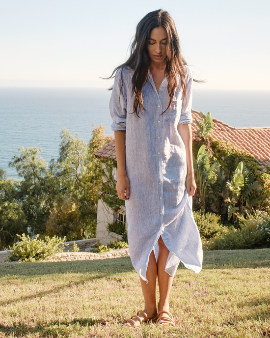A person in a Frank & Eileen Rory Maxi Shirtdress in Blue & White Stripe and sandals standing on grass with the ocean in the background.
