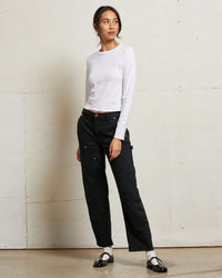 A woman stands indoors against a light wall, wearing the perfectwhitetee Jamie Recycled Cotton L/S in White, black high-waisted cargo pants, and black-and-white Mary Jane shoes. She looks to the side with a relaxed posture.
