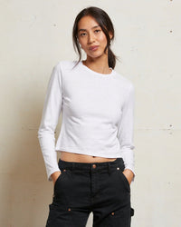 A woman stands against a plain light-colored wall wearing the Jamie Recycled Cotton L/S in White by perfectwhitetee and high-waisted black pants, her left hand in her pocket and a relaxed expression on her face.