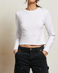 Person wearing the perfectwhitetee Jamie Recycled Cotton L/S in White, paired with high-waisted black pants, standing against a light-colored wall with hands in pockets. Face is not fully visible.