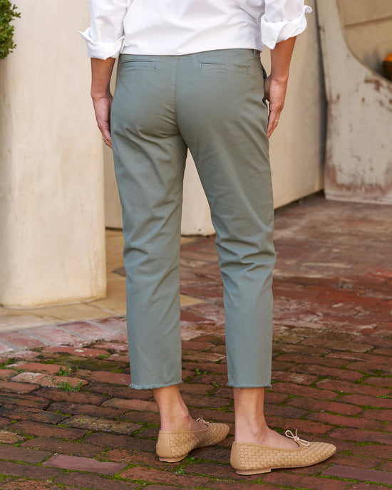 A person wearing Frank & Eileen's Wicklow Italian Chino in Rosemary relaxed fit trousers and beige woven loafers stands on a cobblestone path beside a white wall.