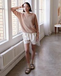 A person standing indoors wearing a beige sweater, white organic cotton Everyday Shorts in Natural from It is well LA, and sandals with socks, posing with one hand on their head.