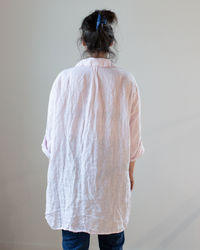 A person seen from behind wearing a loose, crinkled CP Shades Marella Tunic Blouse in Pink Stripe Linen and blue jeans, with their hair tied up using a blue clip.