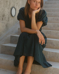 A young woman in the Blair Dress in Sea Pine Check by Trovata Birds of Paradis sits on concrete stairs, resting her chin on her hand as she gazes thoughtfully at the camera. A textured concrete wall with a round light fixture is behind her.