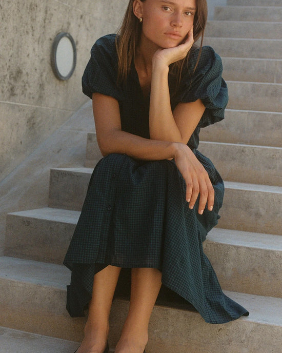 A young woman in the Blair Dress in Sea Pine Check by Trovata Birds of Paradis sits on concrete stairs, resting her chin on her hand as she gazes thoughtfully at the camera. A textured concrete wall with a round light fixture is behind her.