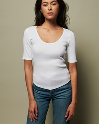 A woman wearing a Nation LTD Lark Deep Scoop Tee in White and blue jeans stands against a neutral background.