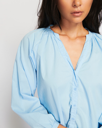 A woman wearing a light blue Nation LTD Desire Dolman Button Up in Shirting peasant blouse stands against a white background, with part of her face cropped out of the frame.