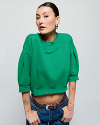 A woman in a Nation LTD Tate Crewneck Sweatshirt in Verdant Green and blue jeans poses confidently, with her hands on her hips and a focused expression. She has visible tattoos on her arms and torso.