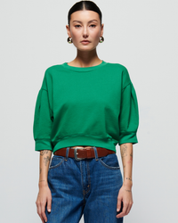 A woman in a Nation LTD Tate Crewneck sweatshirt in Verdant Green and jeans stands confidently against a light background, displaying visible arm tattoos and wearing hoop earrings.