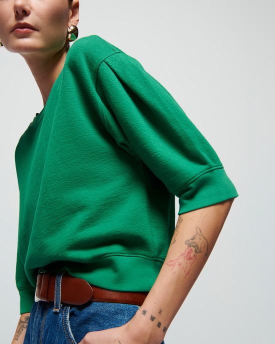 Woman in Nation LTD'S Tate Crewneck Sweatshirt in Verdant Green and jeans with a visible tattoo on her forearm, looking over her shoulder, against a light background.