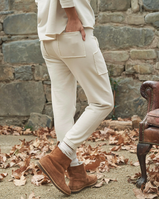 Person standing in a garden wearing Frank & Eileen's Eamon Jogger Sweatpants in Vintage White with brown ankle boots, next to a vintage chair among fallen leaves.