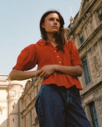 A woman wearing the Trovata Birds of Paradis Jess Blouse in Crimson Check and blue jeans stands outdoors before a historic stone building, looking to the side under a clear blue sky.