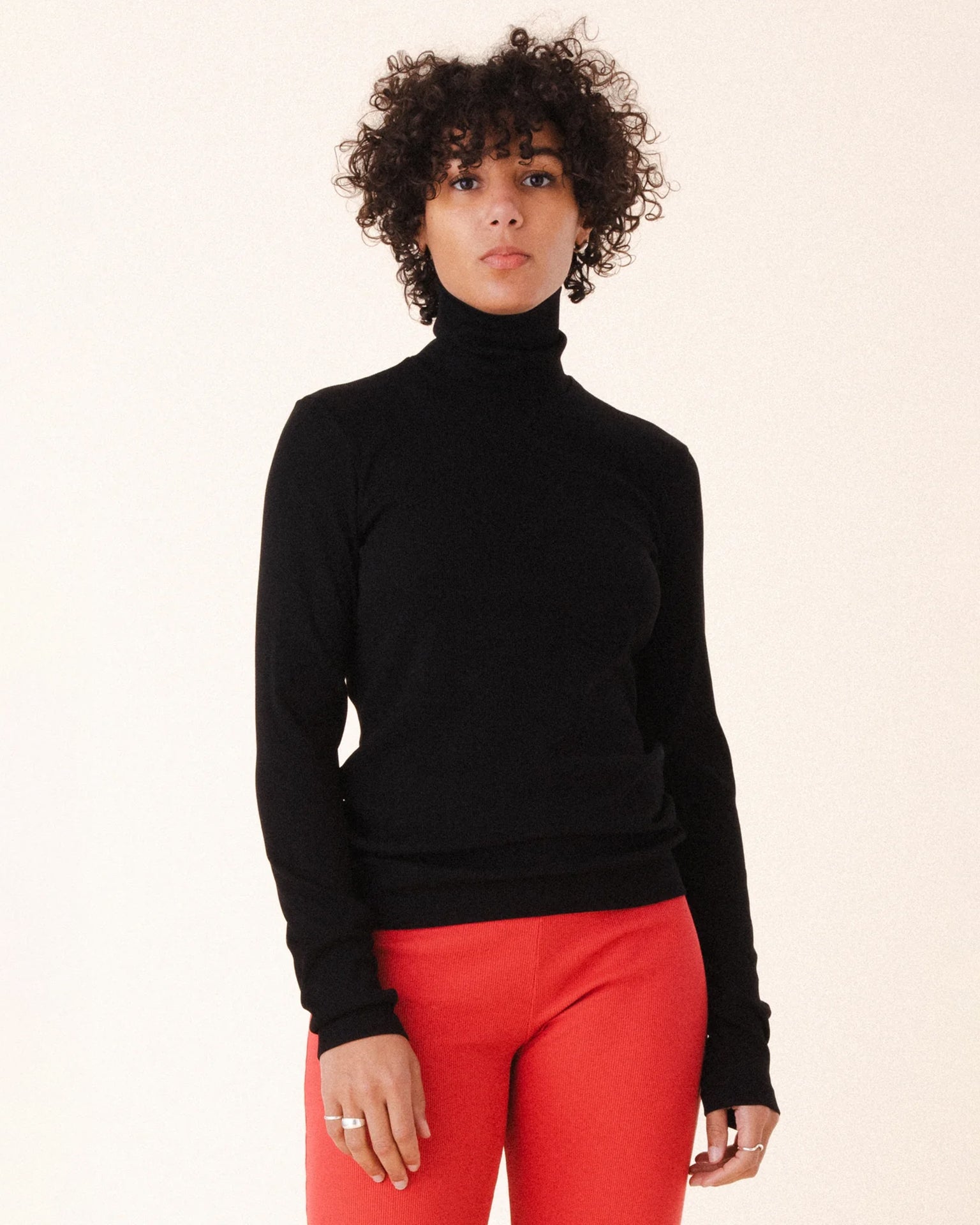 A person with short curly hair wears the Prairie Underground Core Layer in Black, paired with bright red pants, standing against a light background and looking directly at the camera—a stylish, versatile layering piece.