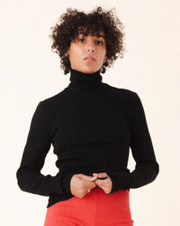 A person with short curly hair wears the Prairie Underground Core Layer in Black with red pants, standing against a plain light background and looking directly at the camera with a neutral expression.