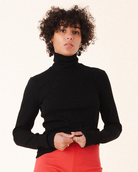 A person with short curly hair wears the Prairie Underground Core Layer in Black with red pants, standing against a plain light background and looking directly at the camera with a neutral expression.