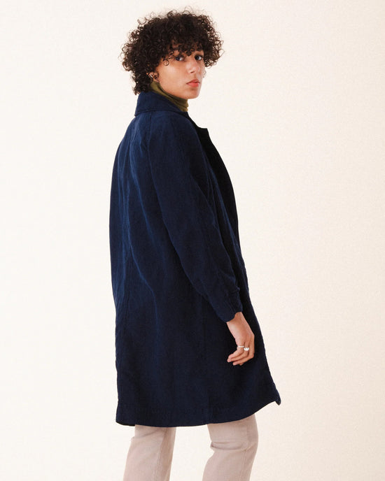 A person with short curly hair stands sideways, looking over their shoulder against a light background, wearing the Prairie Underground Silk Habitual Coat in Midnight over a green turtleneck and light pants.