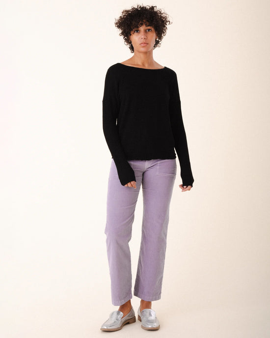 A person with curly hair wears a black long-sleeve top, Prairie Underground Corduroy Carpenter Pant in Silver Mist, and silver loafers, standing against a plain, light background.