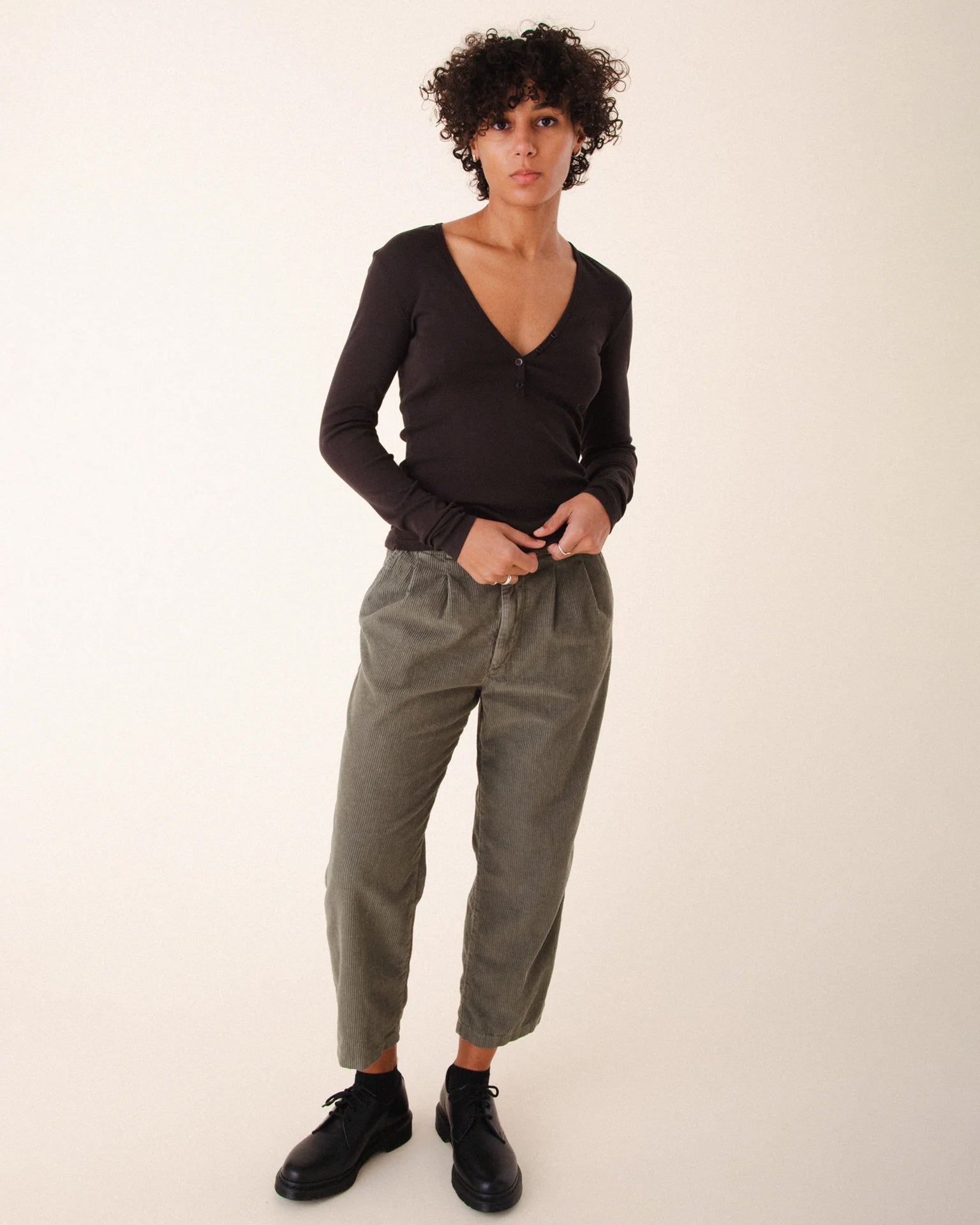 A person with short curly hair wears a fitted dark Prairie Underground V-neck long sleeve top, Prairie Underground Double Pleat Cords in Eucalyptus, and black shoes while standing against a plain light background.