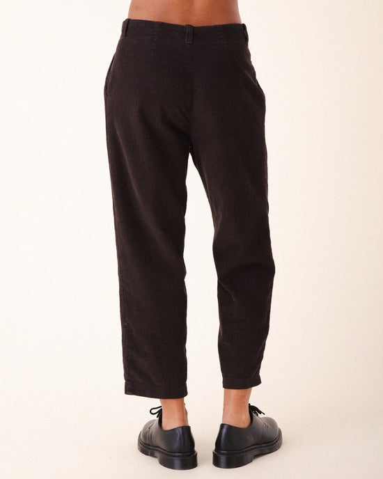 A person wears Prairie Underground's Double Pleat Cords in Brown Out with black oxford shoes, shown from behind against a plain light background. The pants' slightly cropped fit adds vintage flair by revealing the ankles.