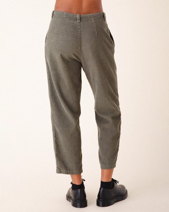 A person stands facing away, wearing Prairie Underground Double Pleat Cords in Eucalyptus—olive green, wide wale corduroy trousers with side pockets—paired with black socks and shoes. The background is plain and light-colored.
