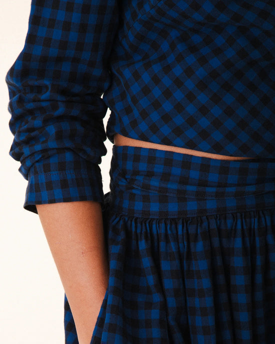 A person wears the Prairie Underground Twist Tie in Blue Mini Check— a long-sleeved, blue and black checkered top with a matching high-waisted skirt, one hand in the skirt pocket. The face is not visible.