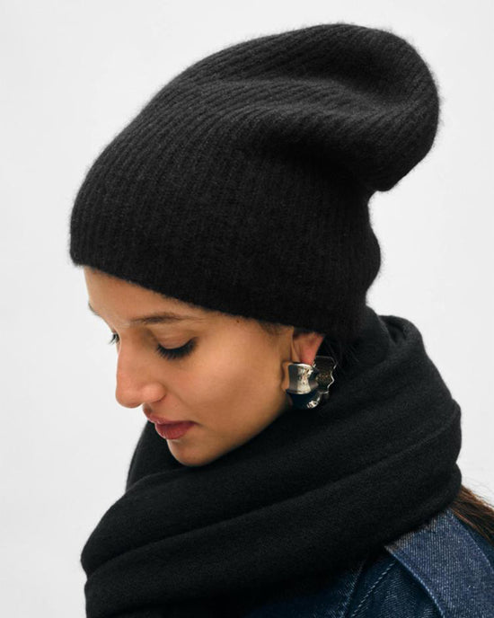 A woman in the White + Warren Cashmere Plush Rib Beanie in Black looks down, styled with a matching scarf, long dark hair, silver earrings, and a dark denim jacket against a light background.